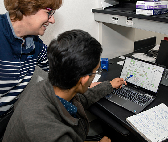 female professor standing behind student sitting in a chair staring at computer screen smiling