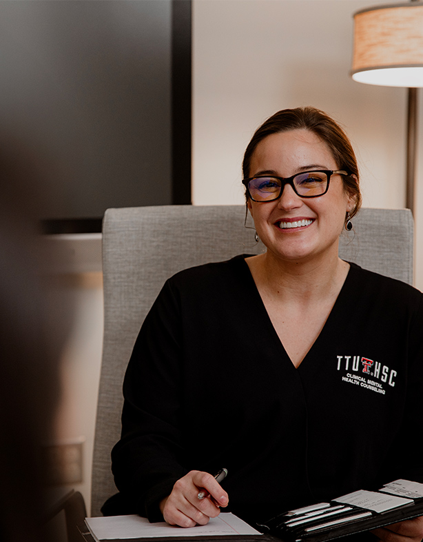 A smiling woman wearing glasses and a black TTUHSC Clinical Mental Health Counseling shirt sits at a desk holding a pen. 