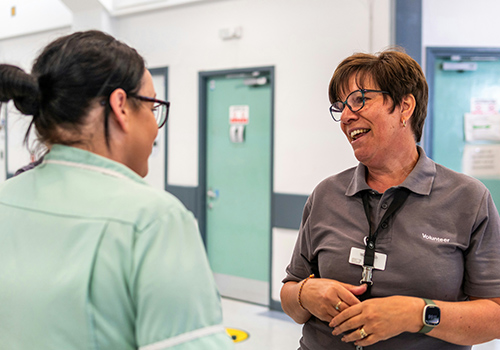 Two people standing and smiling with lanyards around their necks inside a building in conversation