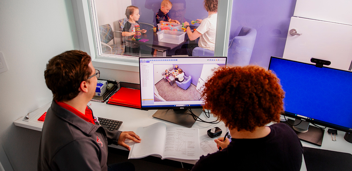 Two TTUHSC providers sitting in a computer room watching children play through a window with another provider