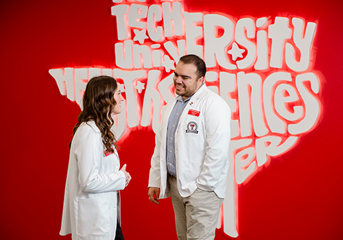 Two TTUHSC students in white lab coats standing infront of the led Texas sign in the library