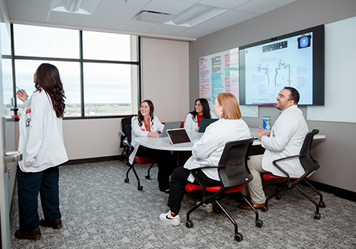 Group of TTUHSC students sitting in a room at a desk while one stands at a dry erase board all with computers open in conversation