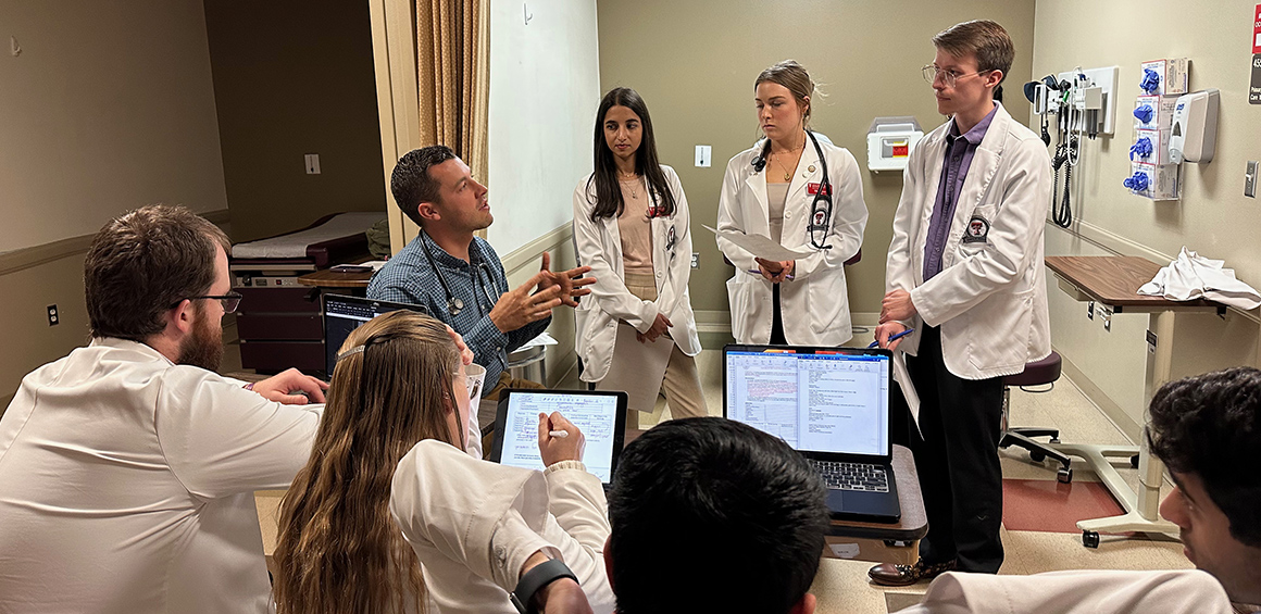 Sevent studnets in white lab coats standing around an instructor who is sitting discussing 