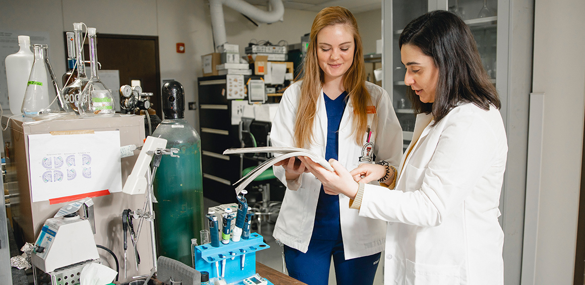 two TTUHSC medical students standing in a laboratory holding a book pointing and smiling