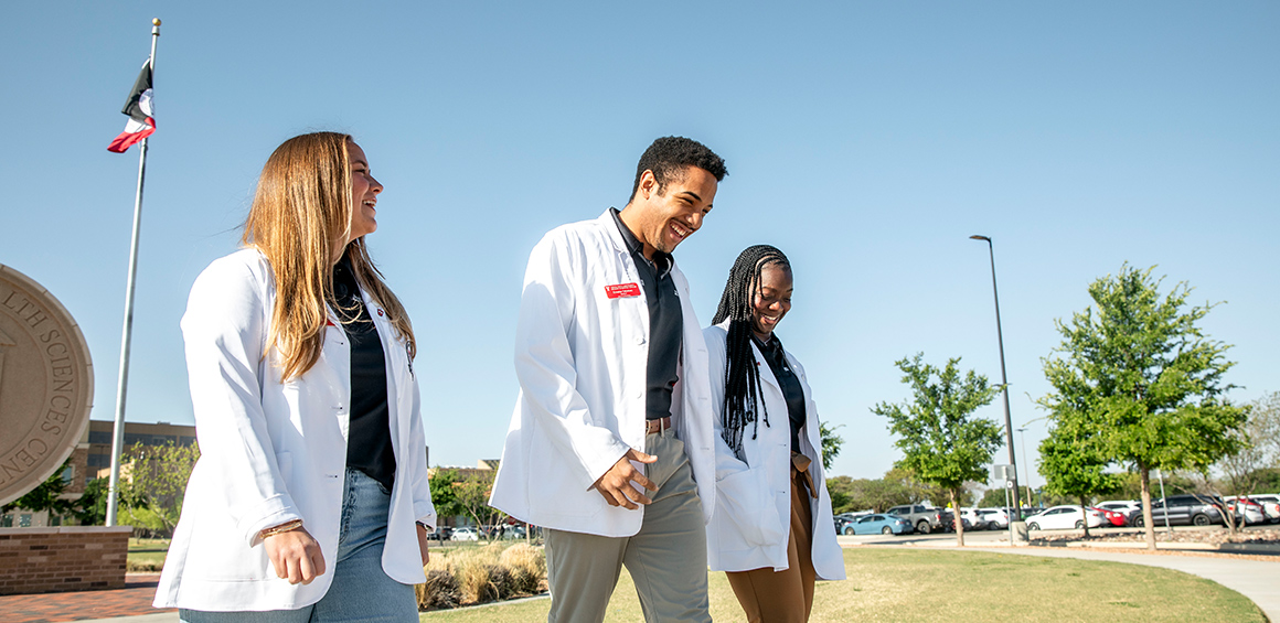 Three TTUHSC students walking in white lab coats smiling with the seal in the background on the Lubbock Campus