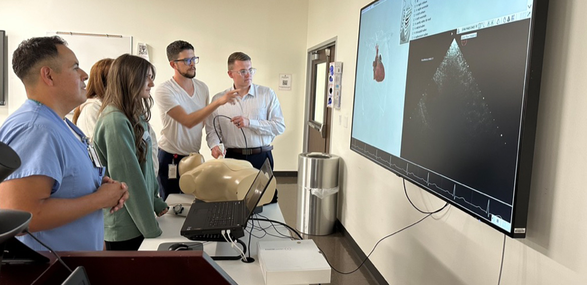 A group of TTUHSC medical students and instructors gather around a mannequin while viewing and discussing ultrasound images displayed on a large screen.