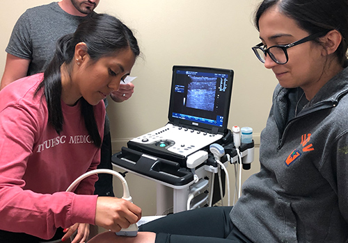 TTUHSC students in a patient room one sitting on a table while the other holds a transducer on the knee of the sitting student