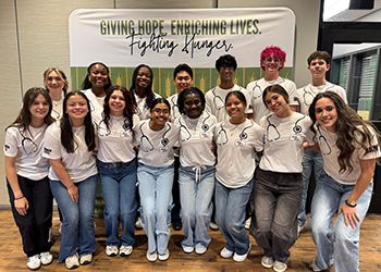 A group of TTUHSC Summer Enrichment students serving at a food bank standing infront of a banner with arms around each other