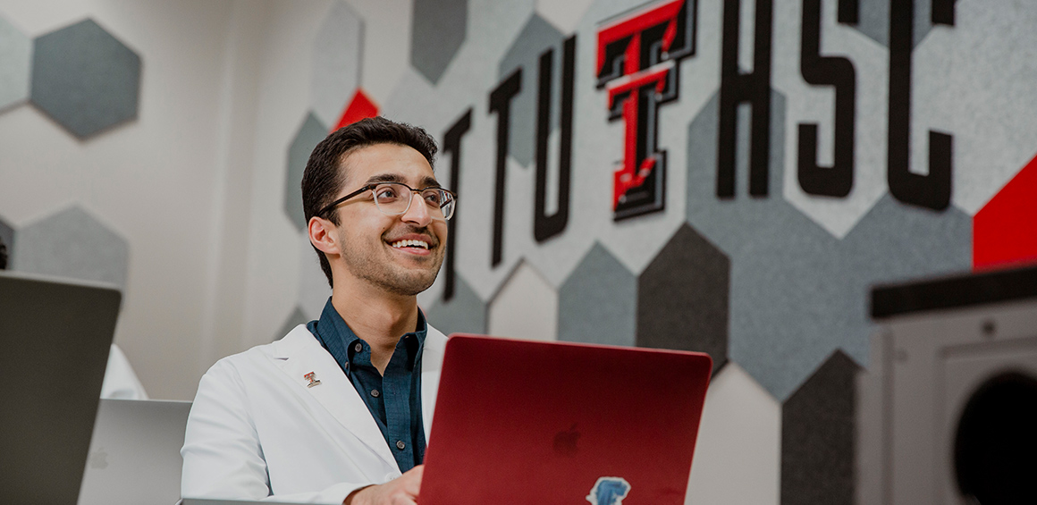 TTUHSC student sitting at a desk in a white lab coat smiling on the computer in a classroom