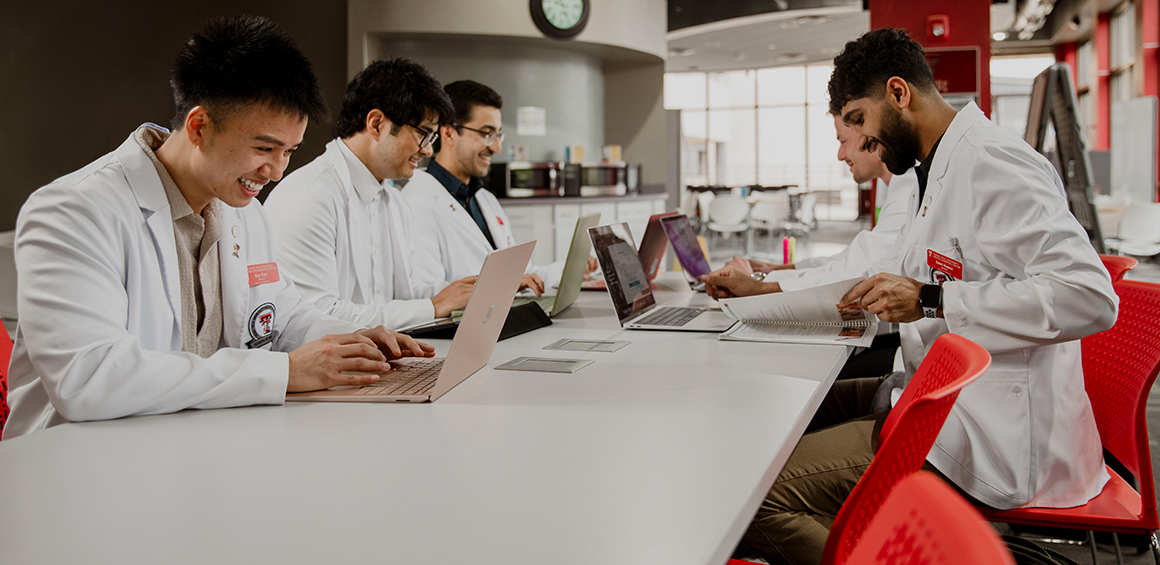 TTUHSC students in white lab coats on laptops smiling sitting around a desk in a lounge area of TTUHSC Lubbock campus