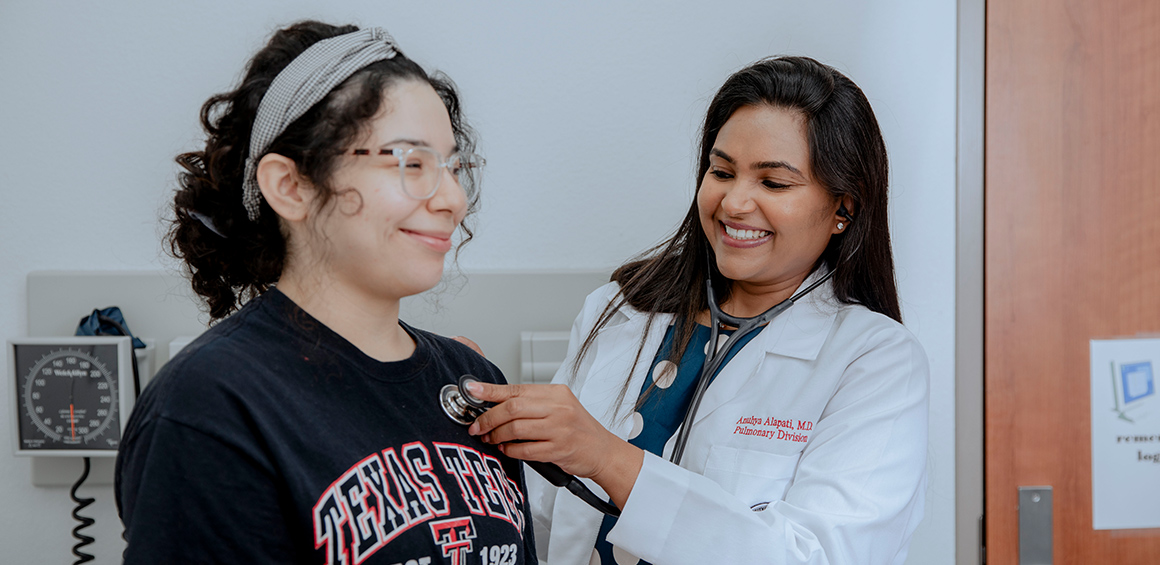 TTUHSC female resident in white lab coat holding stethoscope on a females heart while patient sits on patient table smiling.