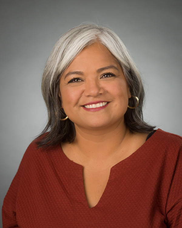 Professional headshot of a smiling person with shoulder-length hair, wearing gold hoop earrings and a rust-colored blouse against a gray studio background.