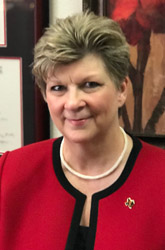 Professional headshot of a smiling person with short brown hair, wearing a red blazer with black trim and a small lapel pin.