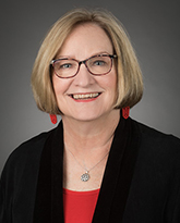 Professional headshot of a smiling person with a blonde bob haircut, glasses, red earrings, and a black top against a gray background.