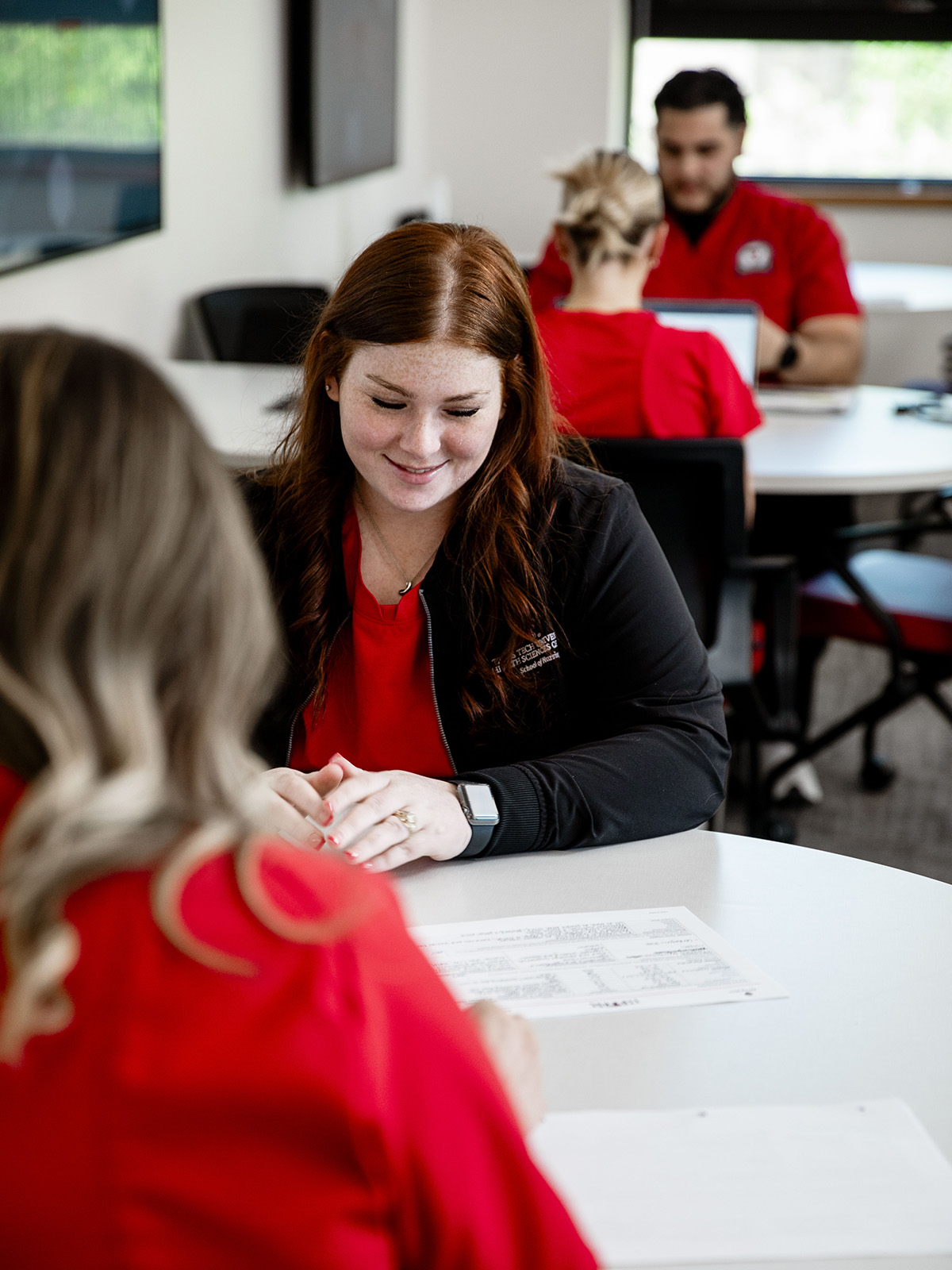 TTUHSC Amarillo students wearing red scrubs sitting together at tables studying looking at a paper.