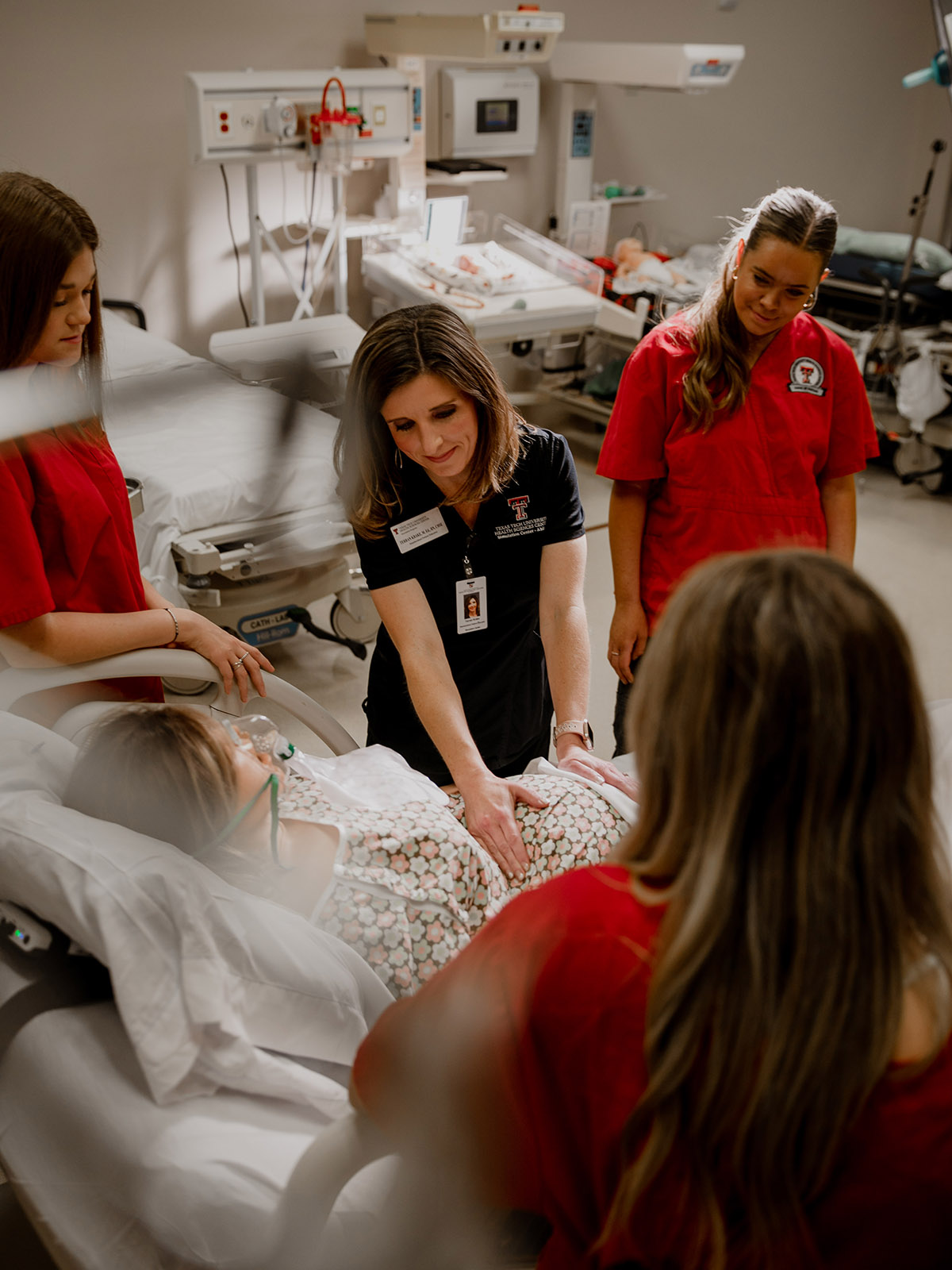 Instructor touching pregnant belly of mannequin patient sitting on a hospital bed with students standing around wearing red scrubs.