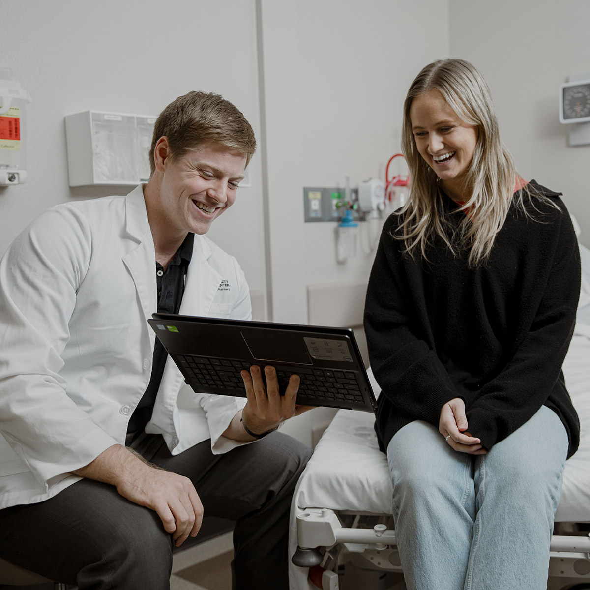 One person sitting on exam table smiling looking at a screen held by person wearing a lab coat in a simulation exam room.