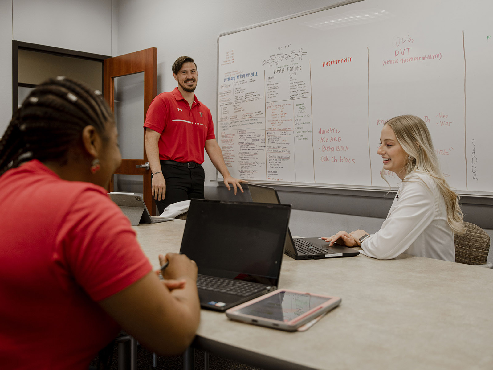 Three TTUHSC Abilene students in a study room writing notes on a whiteboard while looking at their laptops sitting on a table.