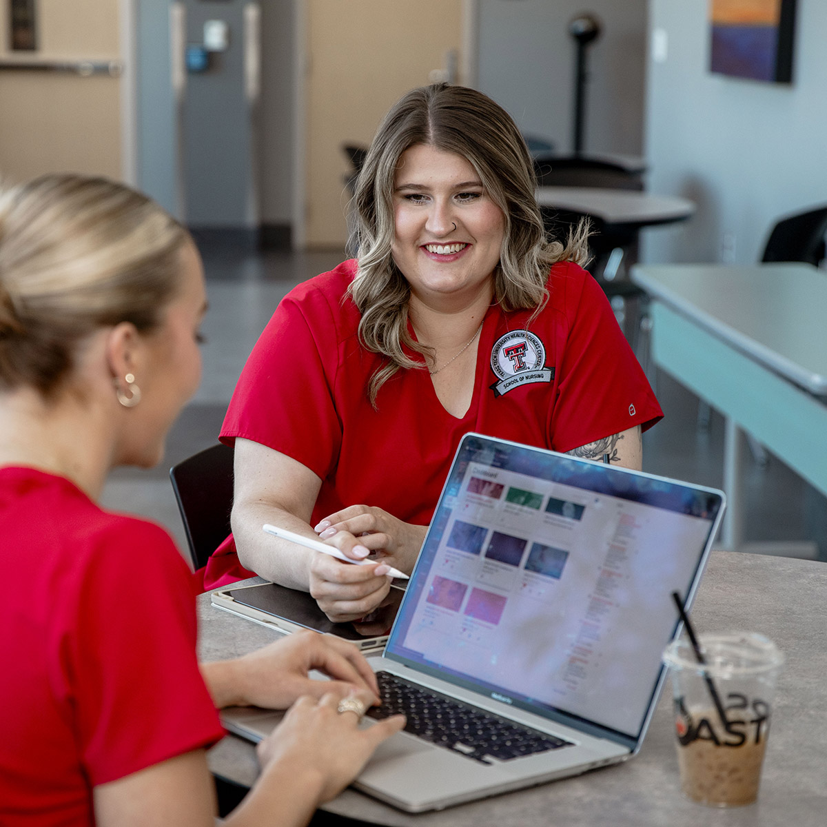 TTUHSC Amarillo students sitting together at a table studying wearing red scrubs looking at screens smiling.