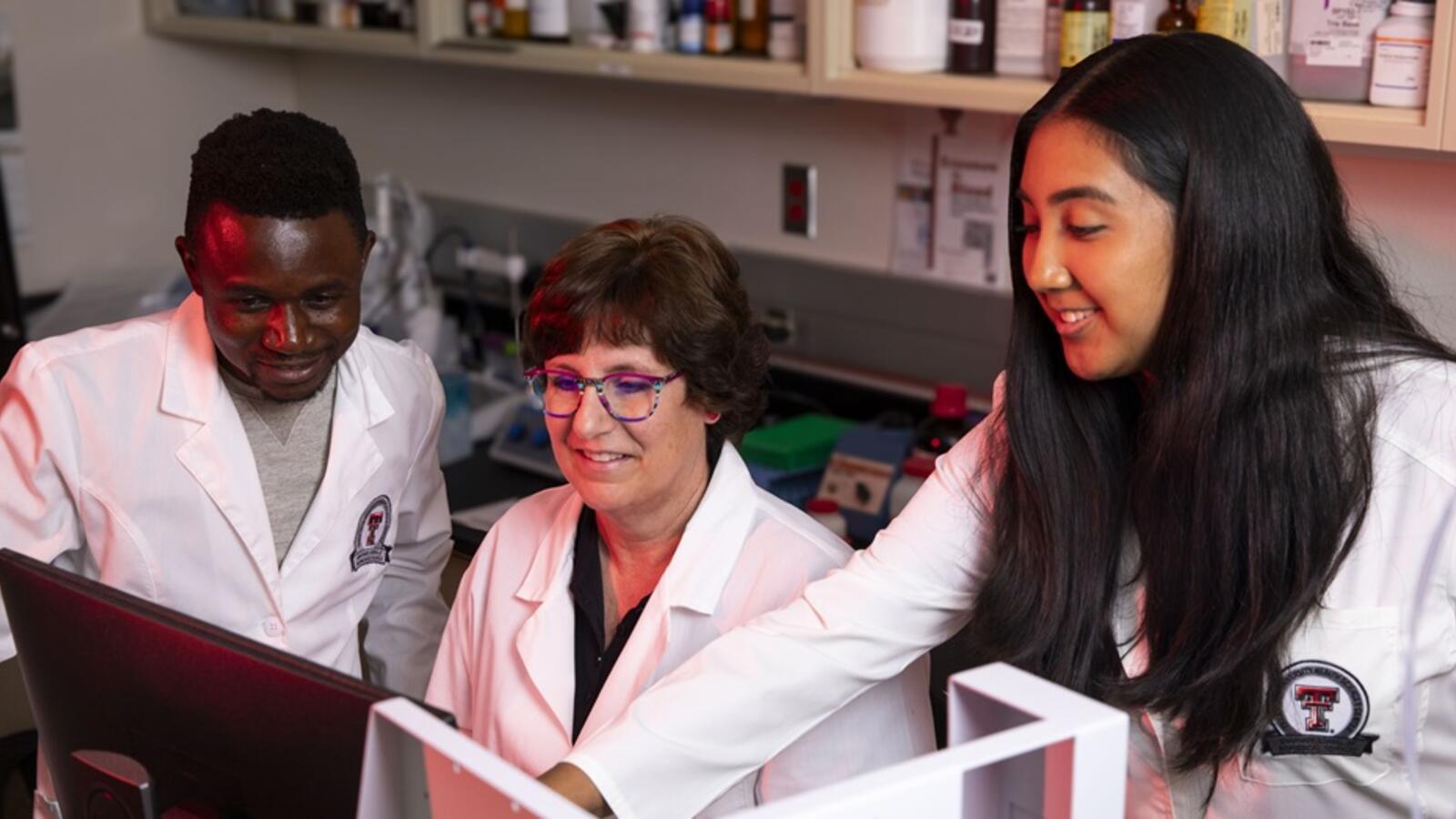 Two students and a professor in white lab coats work in a lab together