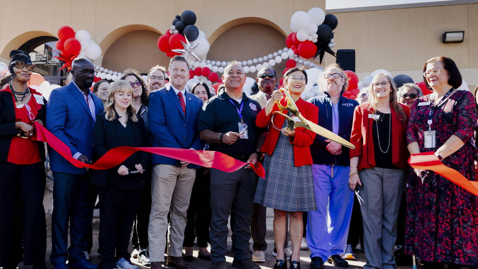 Many people gather outside the Grand Expectations Health Center to cut a ceremonial red ribbon with a large pair of scissors.