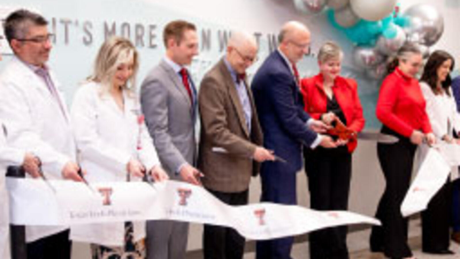 Long line of doctors and administrators cut a ribbon with a large pair of scissors.