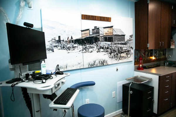 Examination room at the Fort Davis Clinic featuring a mobile medical computer cart, cabinetry, and a large mural titled "FRONTIER MEDICINE" depicting an Old West town.