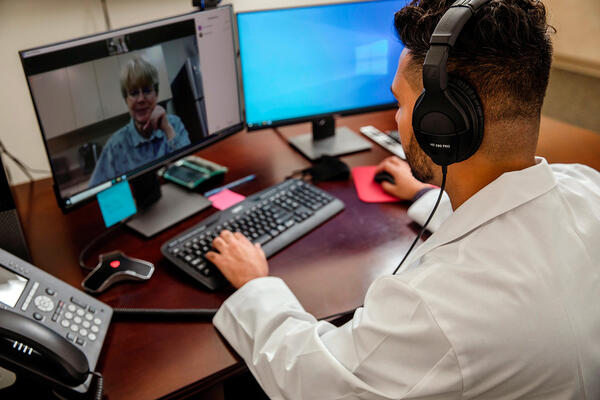 Over-the-shoulder view of a healthcare provider wearing headphones and a lab coat conducting a telehealth video consultation on a computer screen.
