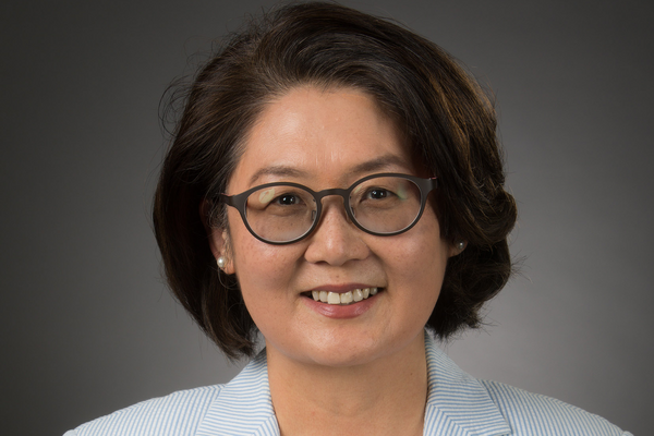 A woman with short brown hair and thin black glasses smiling while wearing a light blue shirt against a grey background.