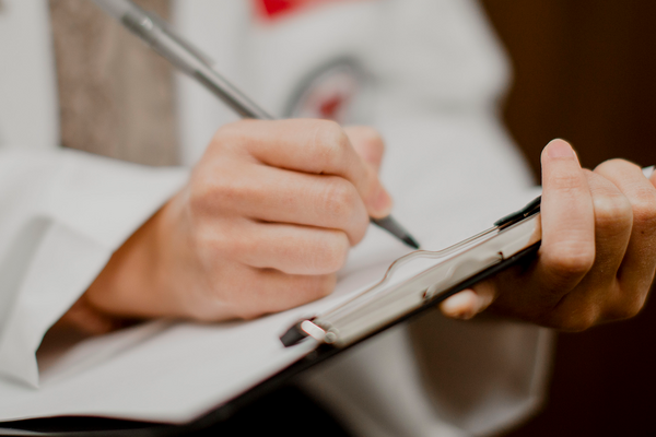 A person wearing a white coat while writing on a clipboard with a grey pen.