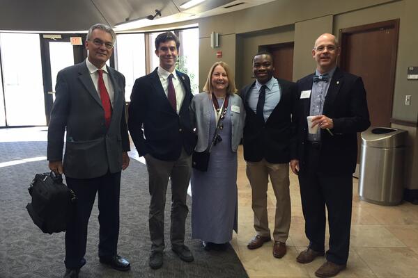 Five professionals in formal attire posing together indoors at an academic or legal event.