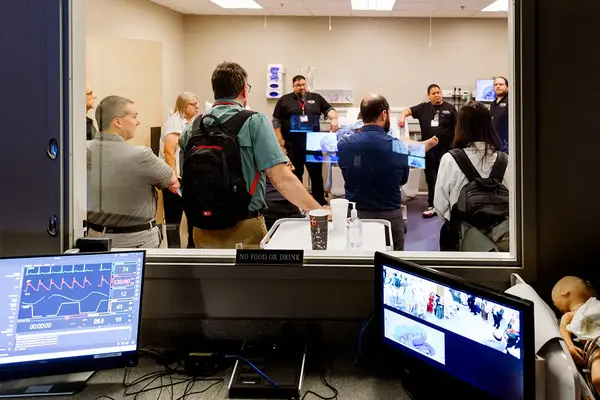 Participants observing a clinical simulation through a control room window with patient monitors