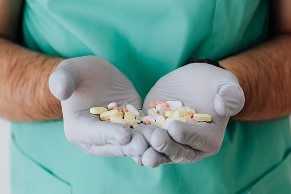 Close-up of a person wearing teal medical scrubs and white gloves holding a handful of assorted yellow and white pills in both hands.