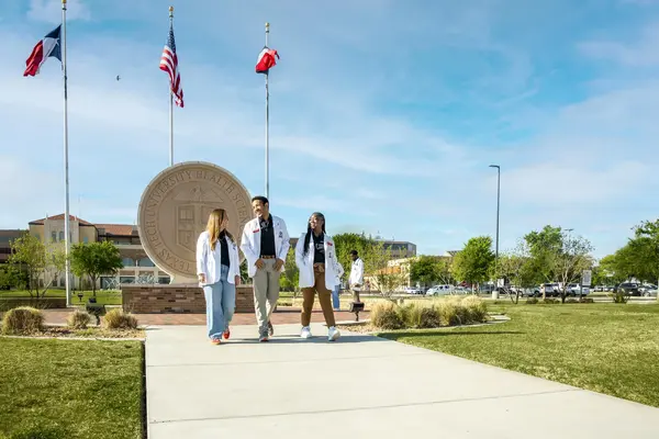 three students in white coats walking away from the university seal sculpture