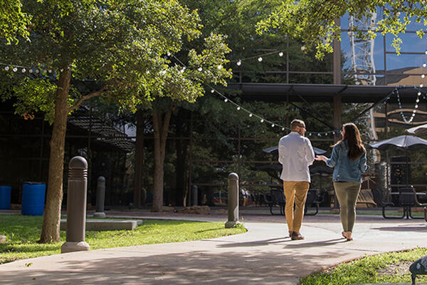 Two people walk along a courtyard path outside a modern TTUHSC building, with trees, patio seating and string lights overhead.
