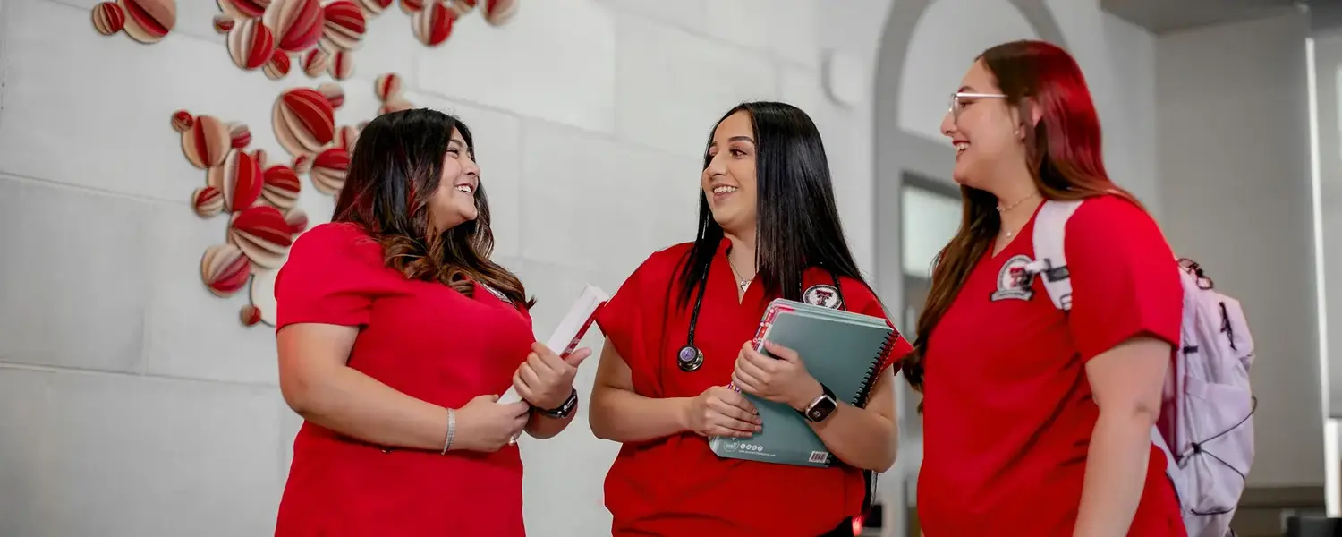 Three healthcare students in red scrubs talking and holding study materials indoors