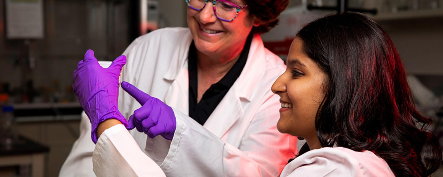 A female professor and a female student in lab coats smiling while looking at a sample vial in a research laboratory.