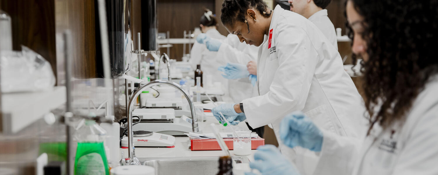 Several people wearing white lab coats and blue gloves are conducting experiments at lab benches in a modern laboratory. The individual in the center is closely focused on handling small lab equipment and containers.