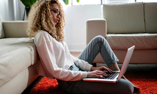 Person seated on the floor using a laptop in a home setting, with a sofa and large windows in the background.