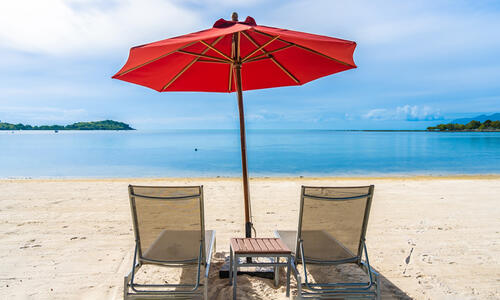 Two beach chairs beneath a red umbrella on a sandy shoreline overlooking calm blue water.
