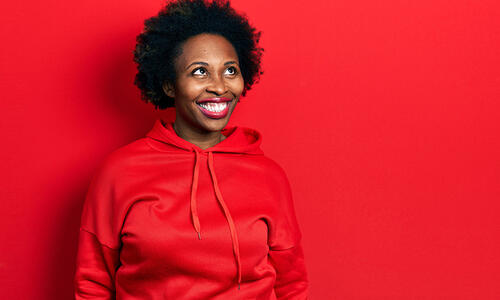 Person wearing a red hoodie against a bright red background, looking upward with a broad smile.