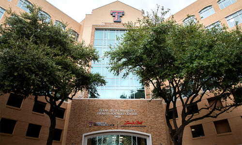 Exterior view of the Texas Tech University Health Sciences Center Dallas campus building, framed by large trees and featuring a glass entrance with TTUHSC signage.