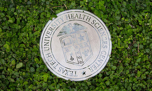Circular metal sign of the TTUHSC seal nestled amongst green shrubery.