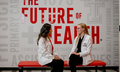 Two TTUHSC students in white coats sit on a red bench in front of a wall graphic displaying the words “The Future of Health.”