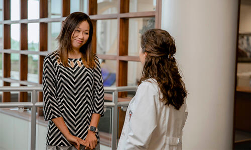Two people converse inside a TTUHSC facility near a windowed corridor, with one person wearing a white clinical coat.