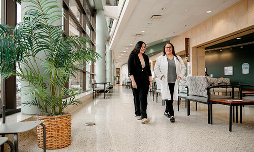 Two TTUHSC team members walk through a bright clinic corridor with large windows, indoor plants and seating areas along the hallway.