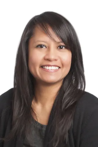 Headshot portrait of a smiling woman with long dark hair, wearing a dark top against a plain white background.
