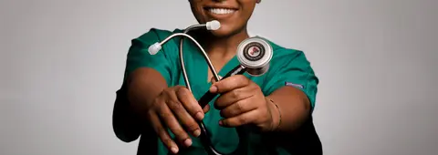 A healthcare worker wearing green scrubs holds out a stethoscope featuring a Texas Tech University Health Sciences Center emblem on the chestpiece.