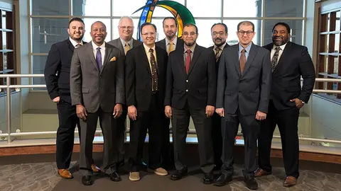 A formal group portrait of nine male healthcare professionals and faculty wearing suits and ties, standing on an upper floor balcony.