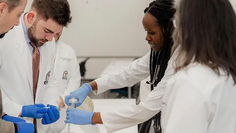 Medical trainees wearing white lab coats and blue gloves practice airway management on a training mannequin in a clinical classroom.